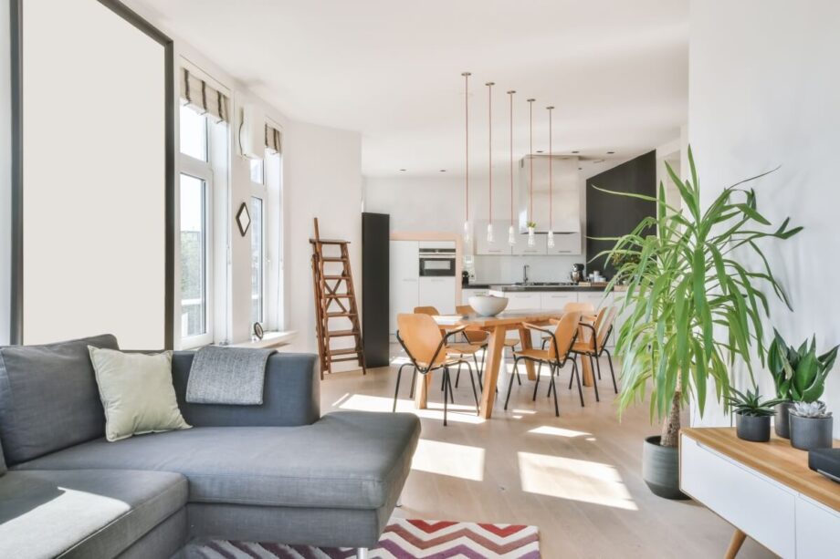 Nowoczesny salon połączony z kuchnią, z jasnymi ścianami, dużymi oknami i roślinami doniczkowymi. Wnętrze ukazuje wpływ technologii smart home w odmianach, podkreślając komfort i funkcjonalność przestrzeni.